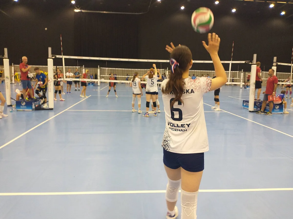 Jugadora joven de espaldas, con el número 6, lanzando el balón con una mano para sacar en un campo de minivoley