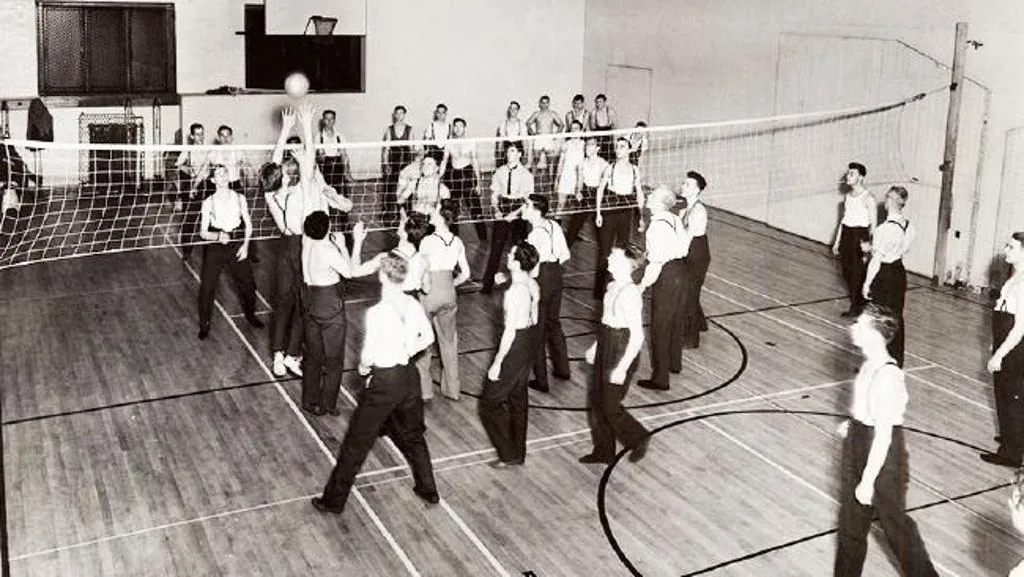 Foto antigua en blanco y negro de un partido de voleibol en un gimnasio, con muchos jugadores a cada lado de la red