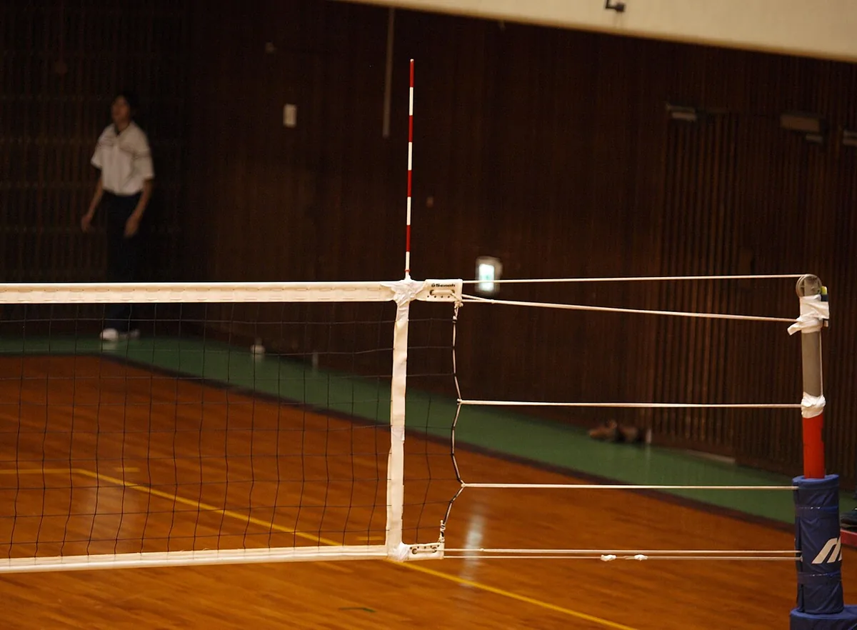 Antena de voleibol con franjas rojas y blancas fijada a la banda lateral de la red, con el poste acolchado al lado